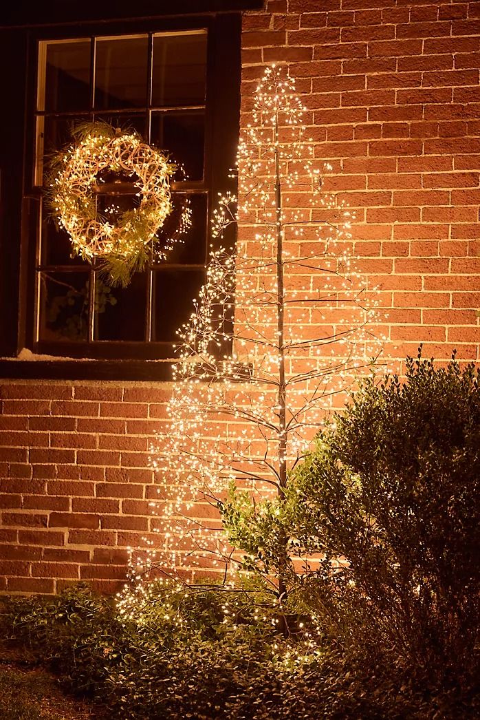 a lighted christmas tree in front of a brick building with a wreath on the window sill