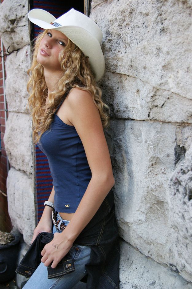 a beautiful young woman wearing a cowboy hat leaning against a stone wall with her hand on her hip