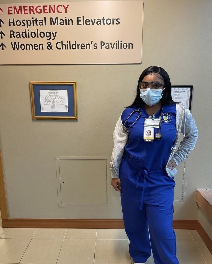 a woman in blue overalls and a face mask standing next to a hospital sign