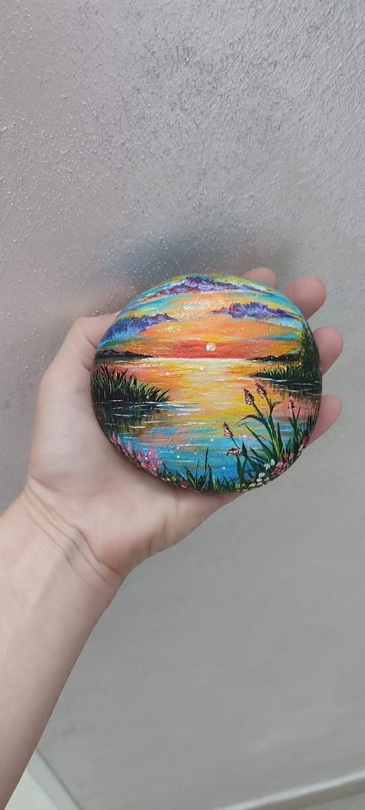 a hand holding a painted rock in front of a white wall with water and trees on it