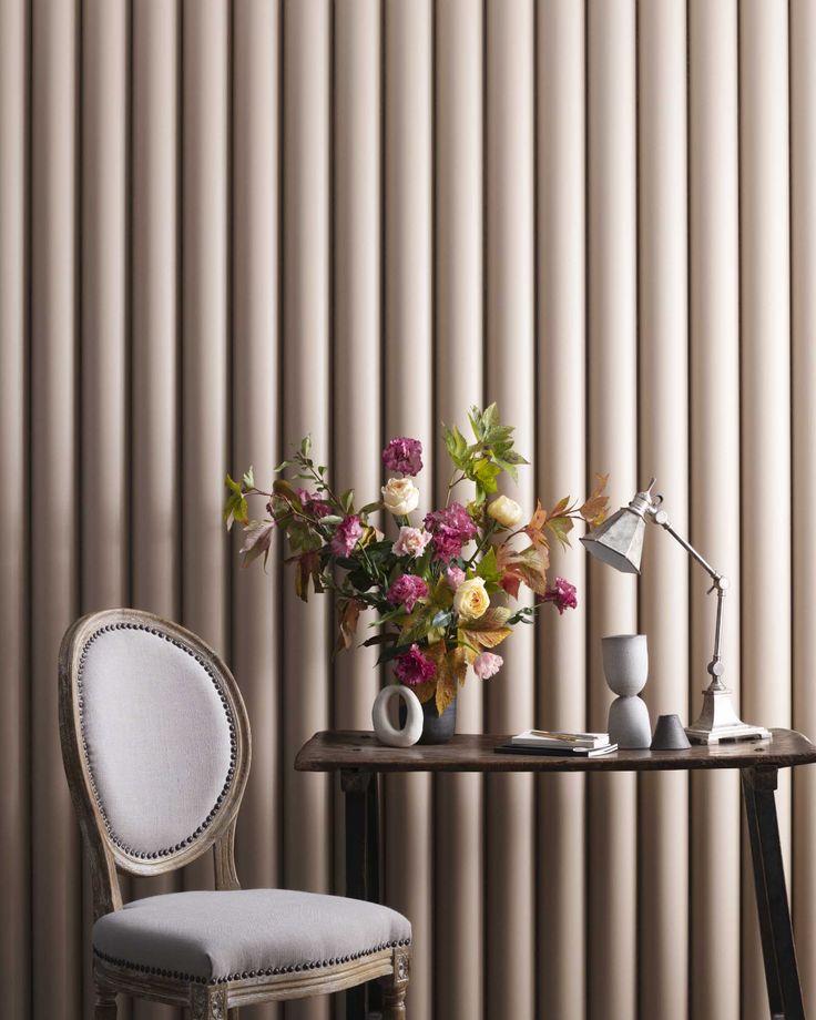 a vase with flowers on top of a table next to a chair in front of a striped wall