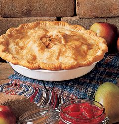 an apple pie sits on a table with apples around it and jars of jam in the background