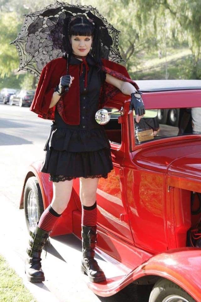 a woman in black and red outfit standing next to an old car with an umbrella over her head