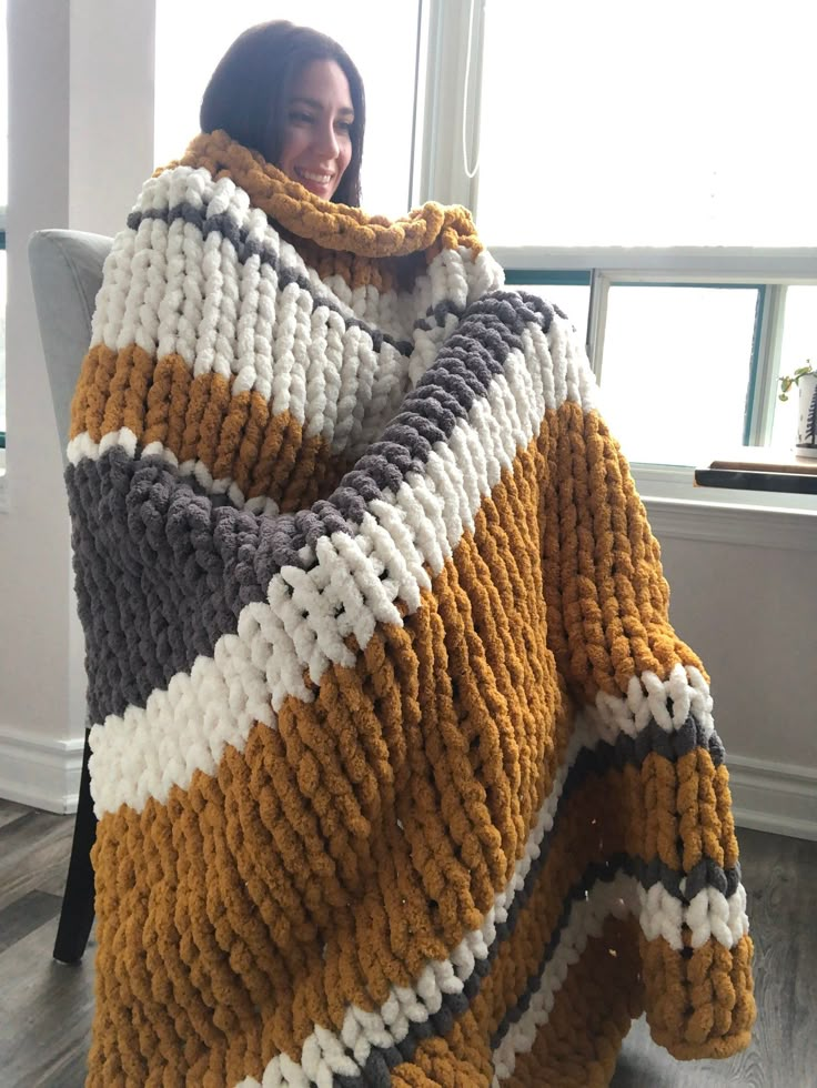 a woman sitting in a chair wrapped up in a large knitted blanket, smiling at the camera