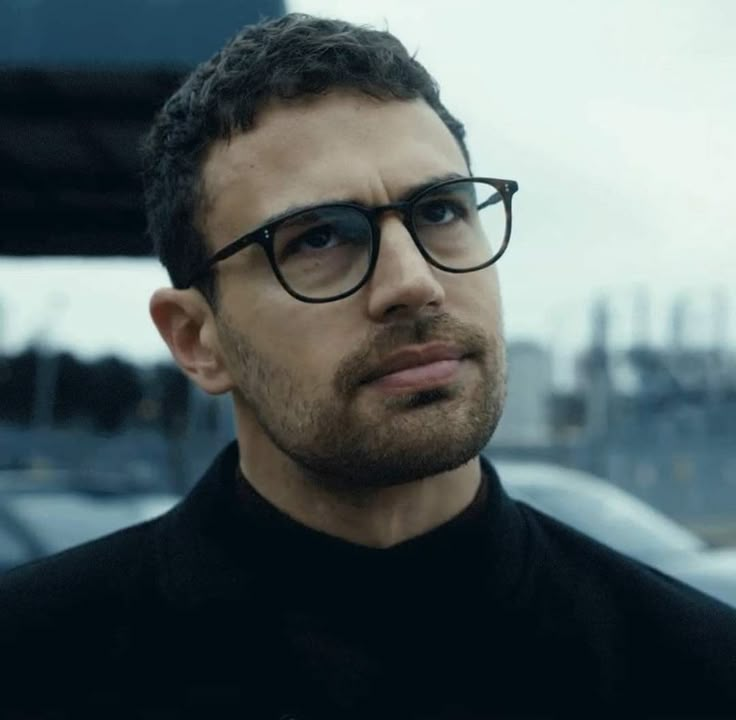 a man wearing glasses standing in front of some cars and looking off into the distance