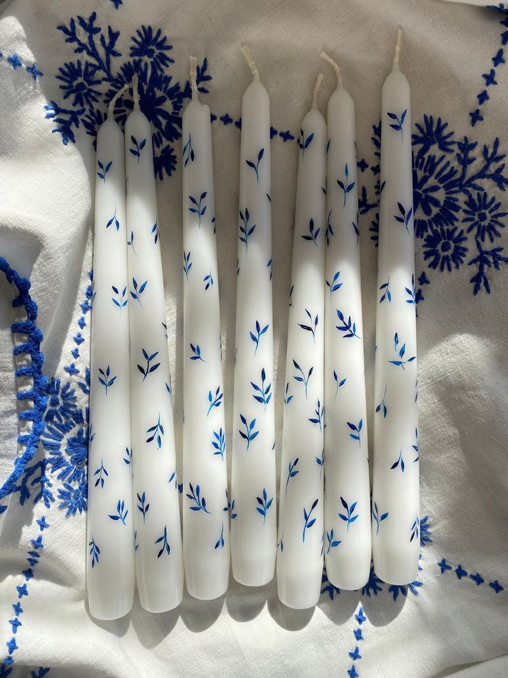 six candles are lined up on a white cloth with blue flowers and leaves painted on them