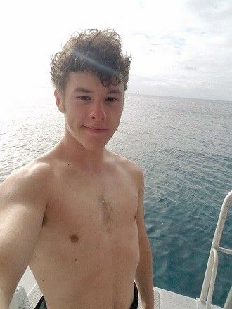 a shirtless young man standing on the back of a boat looking at the camera