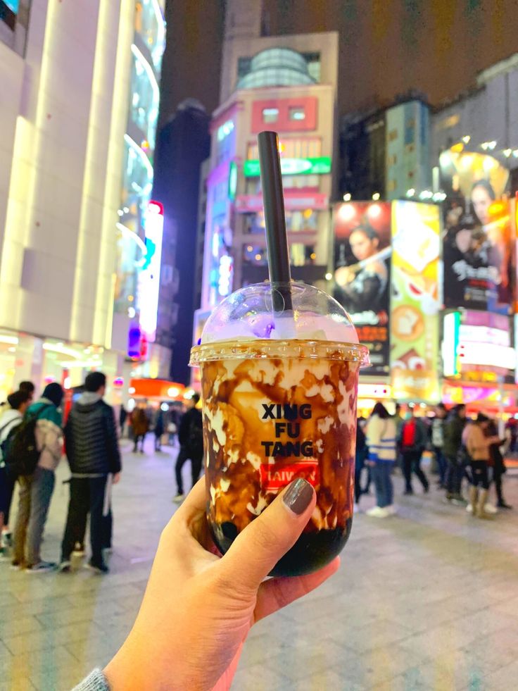 a person holding up a drink in the middle of a busy city street at night