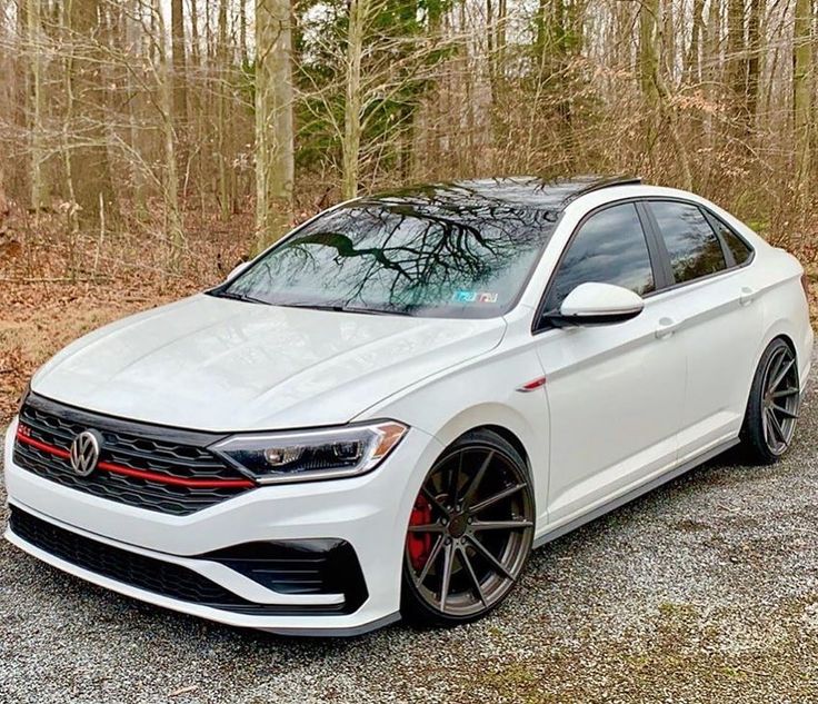 a white volkswagen sedan parked in front of some trees and bushes on a gravel road