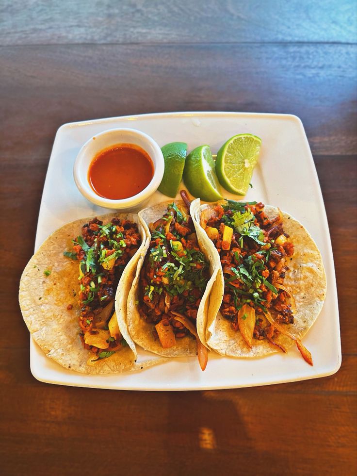 three tacos on a white plate with salsa and lime wedges next to it