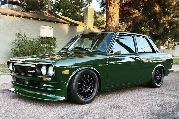 a green car parked on the street in front of a house with trees behind it