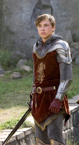 the young man is dressed in medieval armor and holding two swords, standing next to a stone wall