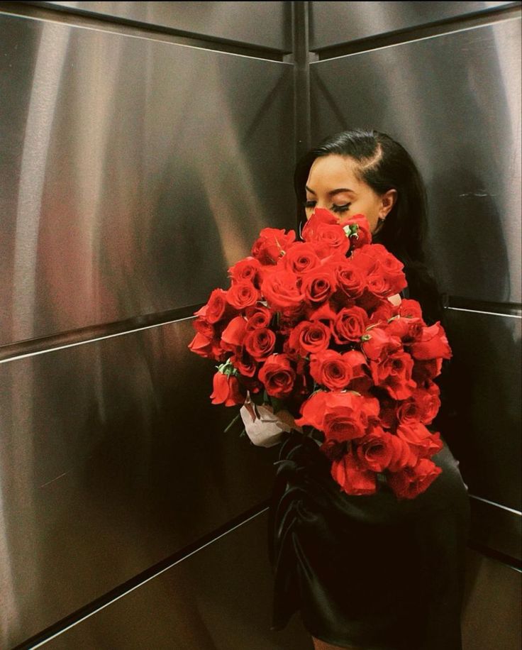 a woman holding a bunch of red roses in front of a metal wall and looking at the camera