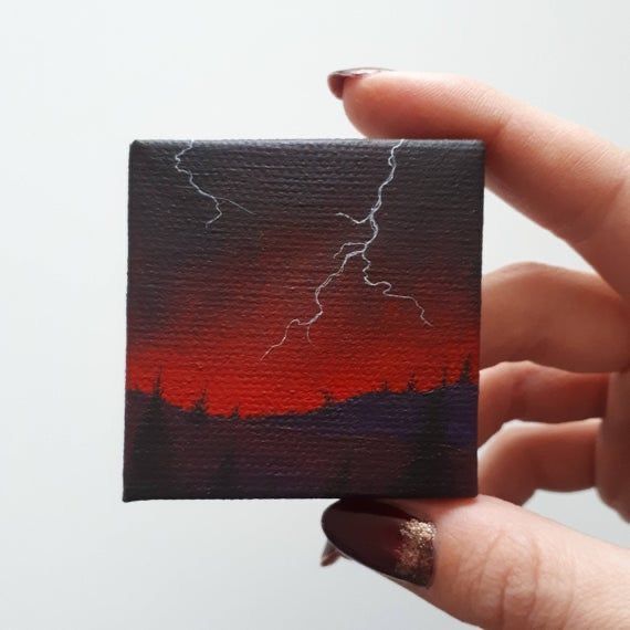 a hand holding up a small square painting with lightning in the sky above it and trees below