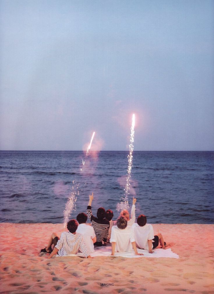 four people sitting on the beach watching fireworks go off in the sky over the water