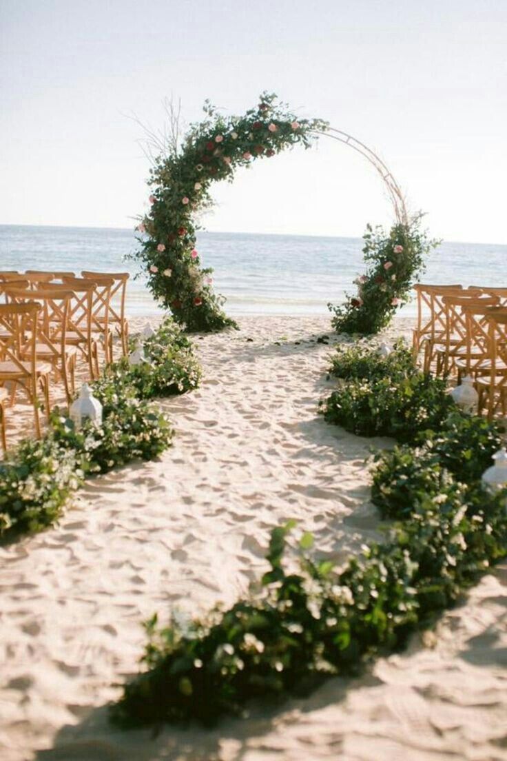 an outdoor ceremony set up on the beach with wooden chairs and greenery in the sand