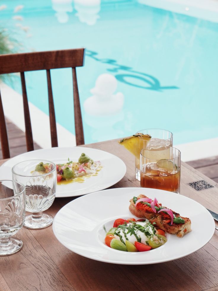 two white plates with food on them next to a swimming pool and table set for dinner