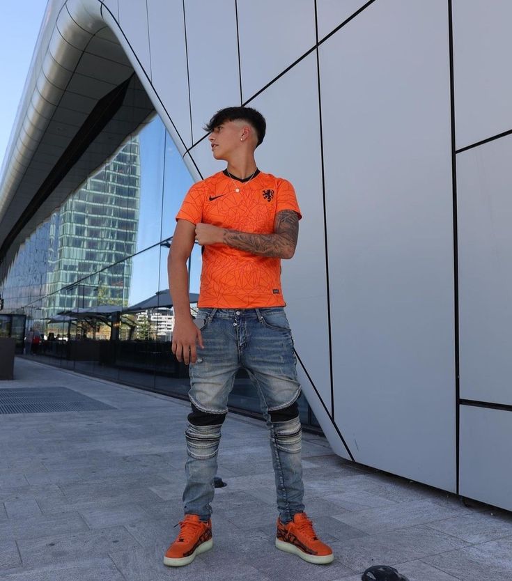 a man in an orange shirt and jeans standing next to a wall with his arms crossed