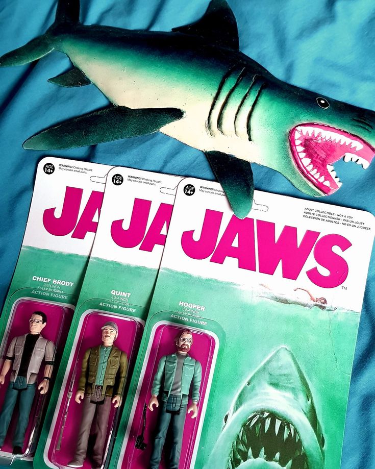 three action figures are displayed on a bed with a shark and man in the background