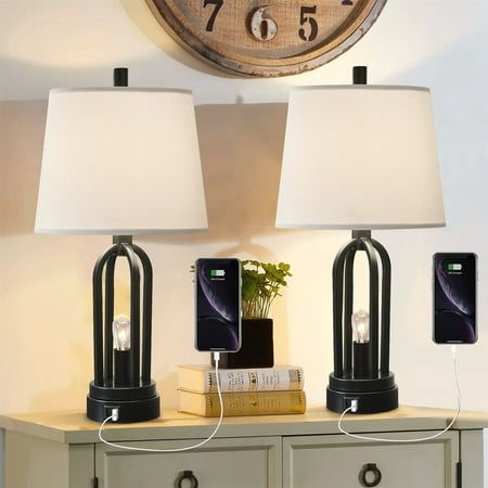 two lamps sitting on top of a wooden table next to a wall mounted clock with roman numerals