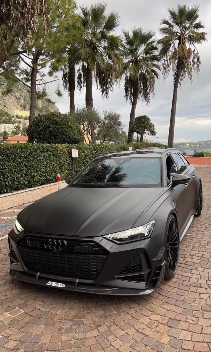 a black sports car parked in front of some palm trees and bushes on a cobblestone driveway