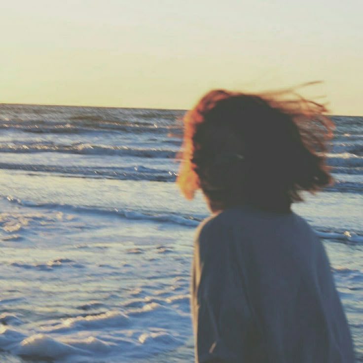 a woman standing on top of a beach next to the ocean with her hair blowing in the wind