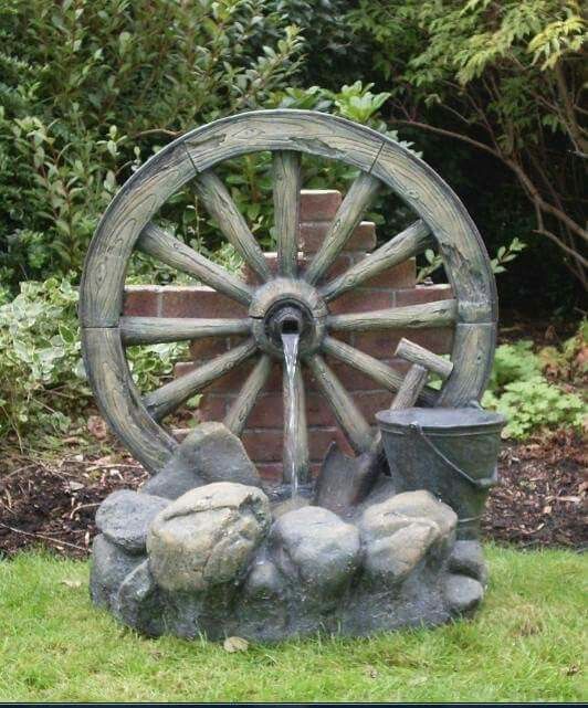 a water wheel with rocks and buckets in front of it on the grass next to a brick wall