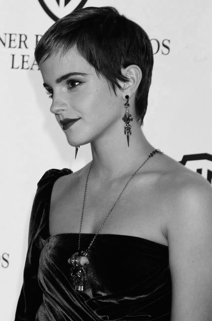 a woman with short hair wearing a black dress and earrings on her neck, standing in front of a wall