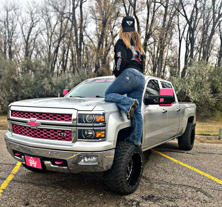 two women are sitting on the hood of a silver pickup truck in a parking lot