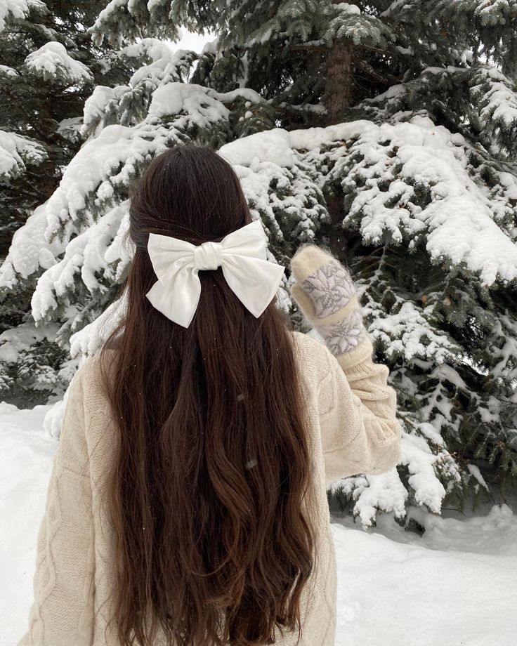 a woman with long hair wearing a white bow in front of snow covered pine trees