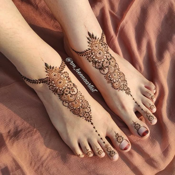 two feet with henna tattoos on them sitting on top of a pink bed sheet