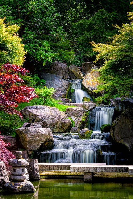 a small waterfall in the middle of a pond surrounded by rocks and trees with red leaves