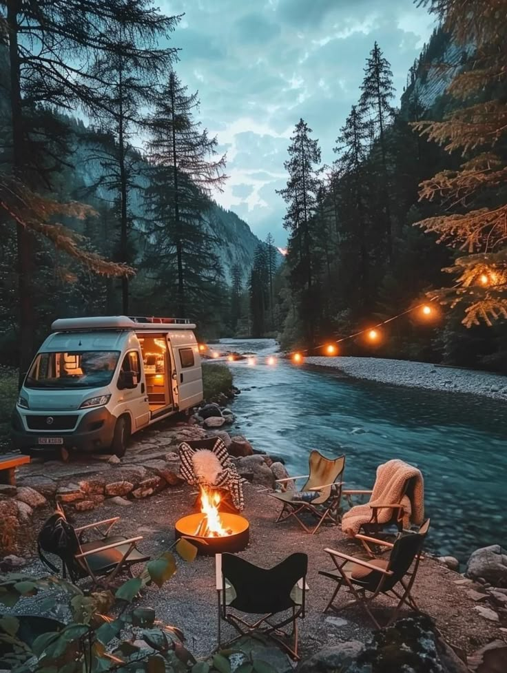 a camper van parked next to a river with chairs and fire pit in the foreground