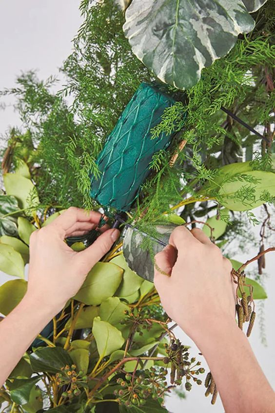 two hands are trimming the branches of a tree with green leaves and artificial greenery