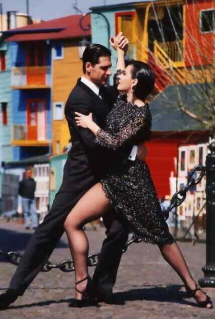 a man and woman dancing in front of colorful buildings on the street with their arms around each other