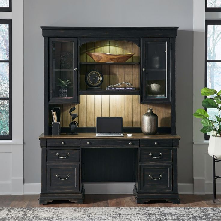 a desk with a laptop on it in front of two windows and a potted plant