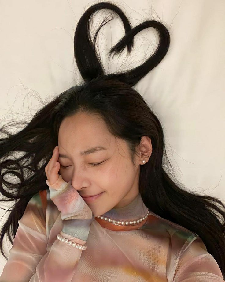 a woman with long black hair laying on top of a white bed next to a heart shaped object