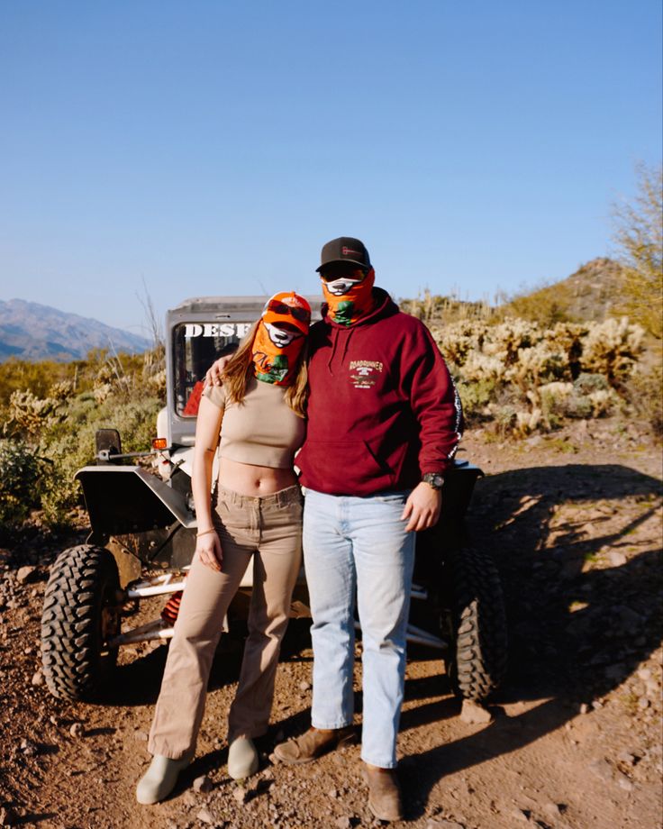 a man and woman standing next to each other in front of a vehicle on a dirt road