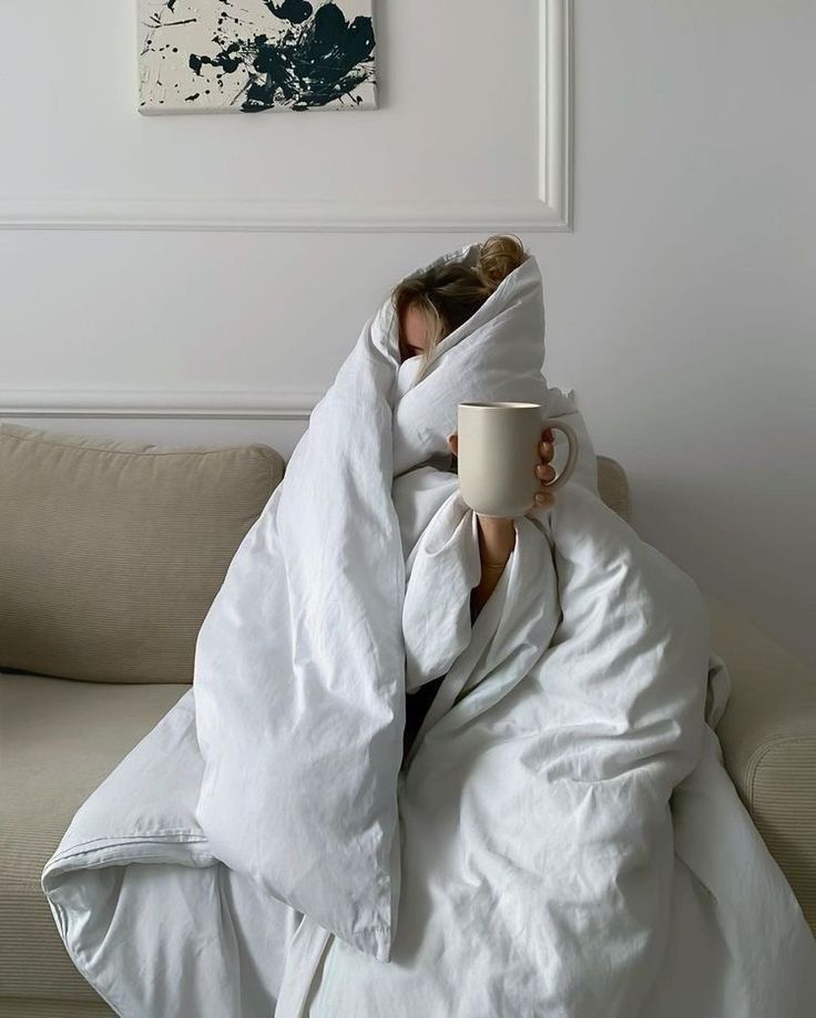 a woman wrapped up in a blanket and holding a coffee cup under her arm while sitting on a couch