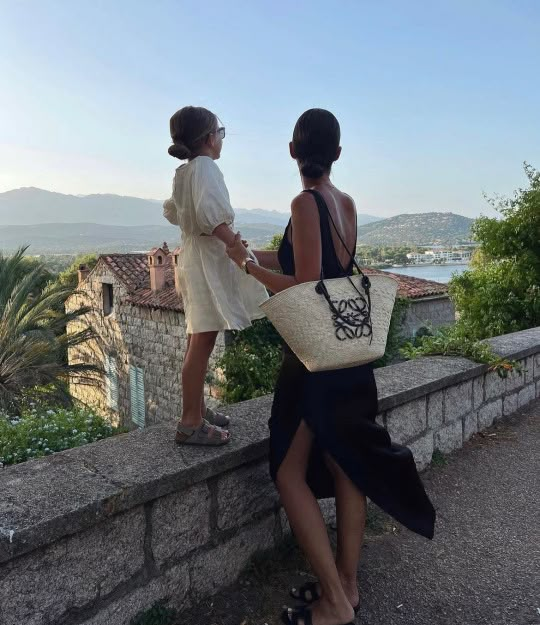 two women standing next to each other near a stone wall and looking at the water