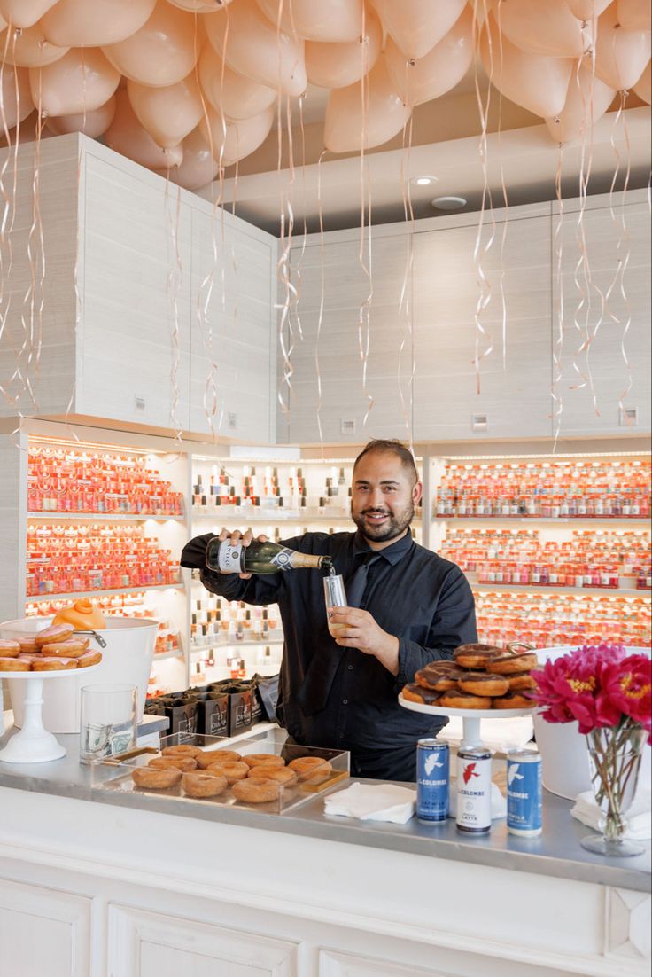 a man pours champagne at a donut shop counter with balloons in the background