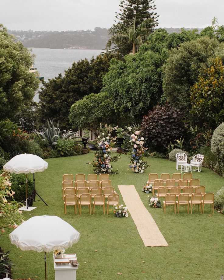 an outdoor ceremony setup with chairs, umbrellas and flowers on the grass near water