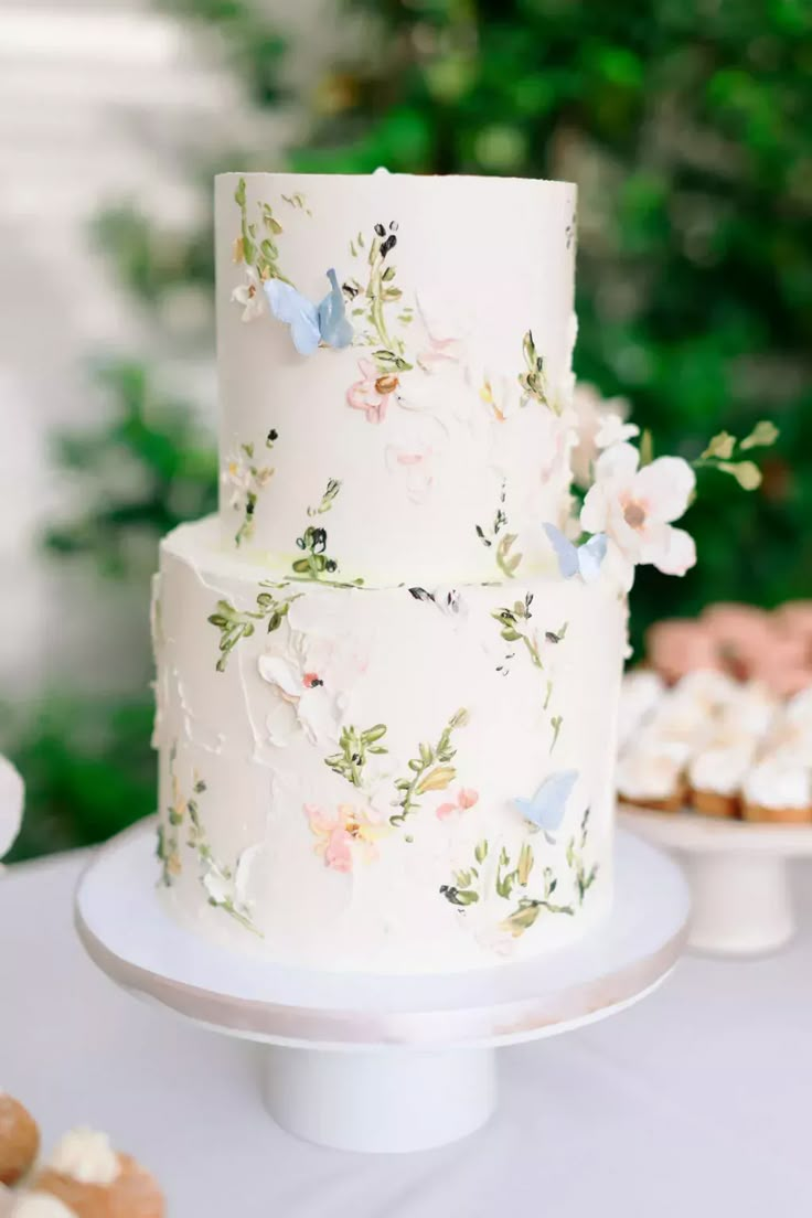 a three tiered white cake with flowers and butterflies on the top is surrounded by cookies