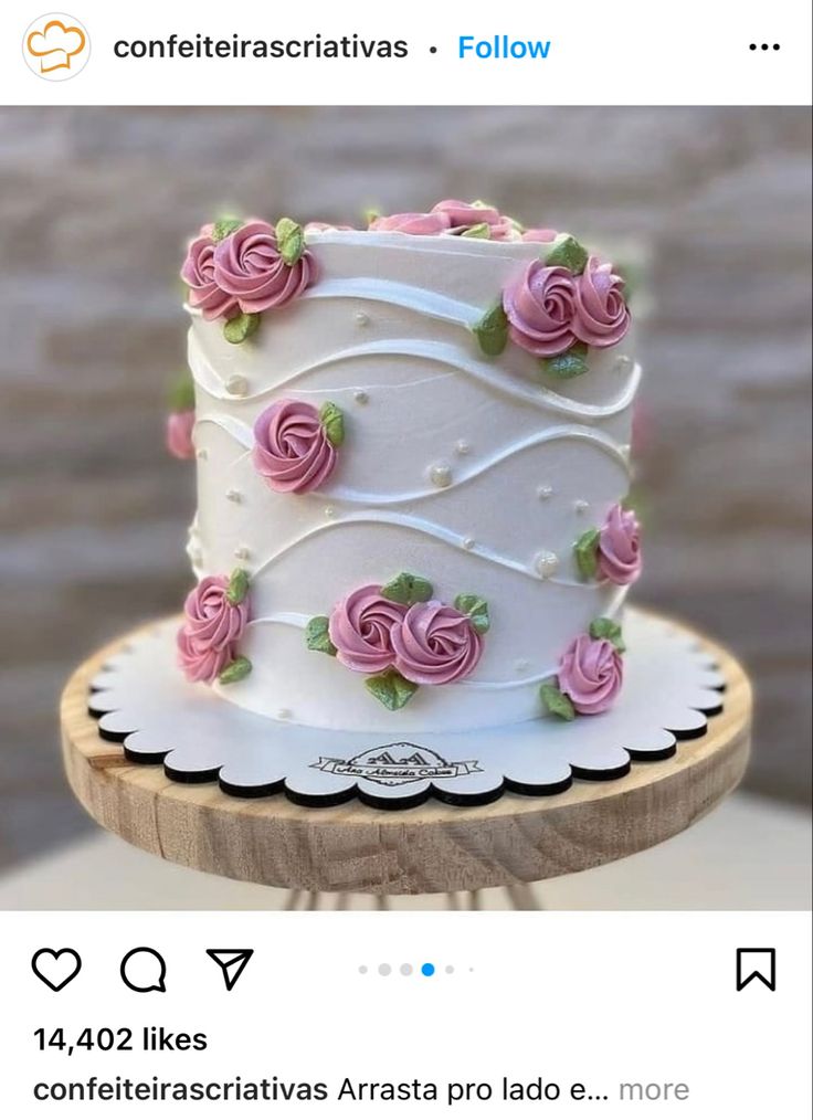 a white cake with pink roses on it sitting on top of a wooden stand next to a brick wall