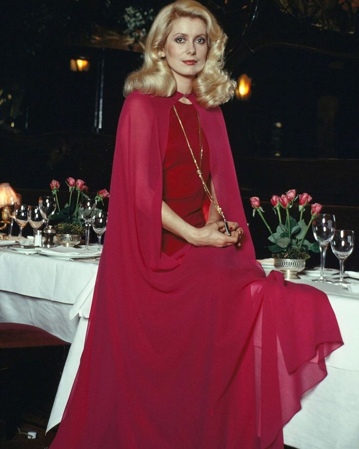 a woman in a red dress and cape sitting at a table with wine glasses on it