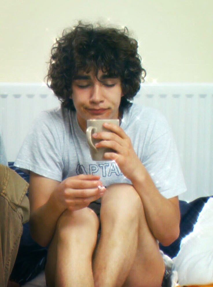 a young man sitting on a bed holding a cup and looking at his cell phone