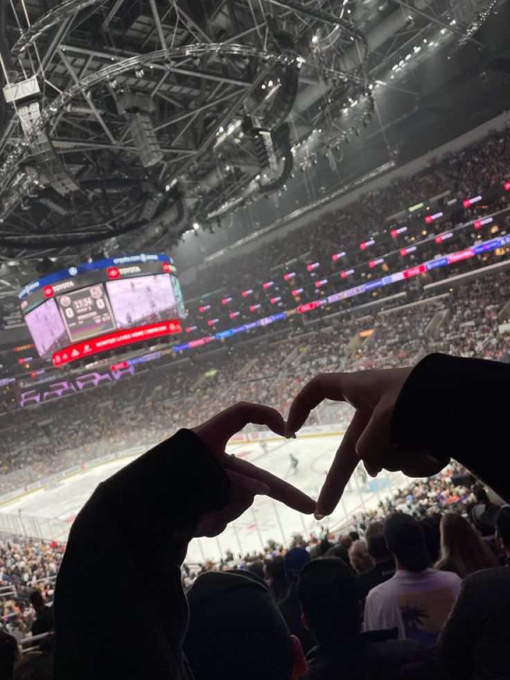 two hands making a heart shape in front of an audience at a hockey game with the scoreboard lit up