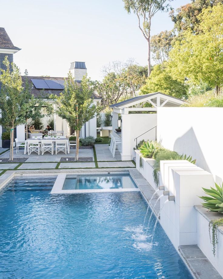 an outdoor swimming pool surrounded by greenery and white chairs, with a dining table in the background