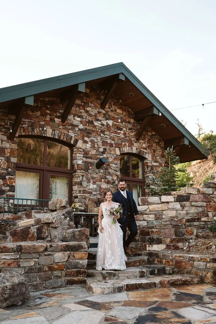 a bride and groom standing on the steps of their stone house in front of it