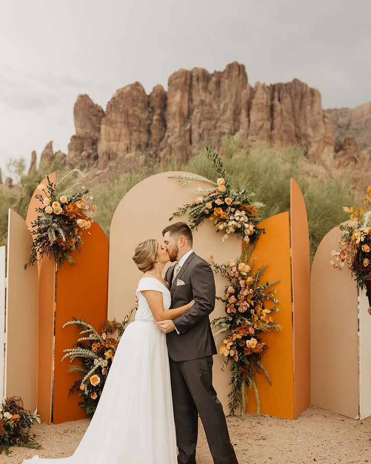 a bride and groom kissing in front of an arch decorated with orange flowers at their wedding